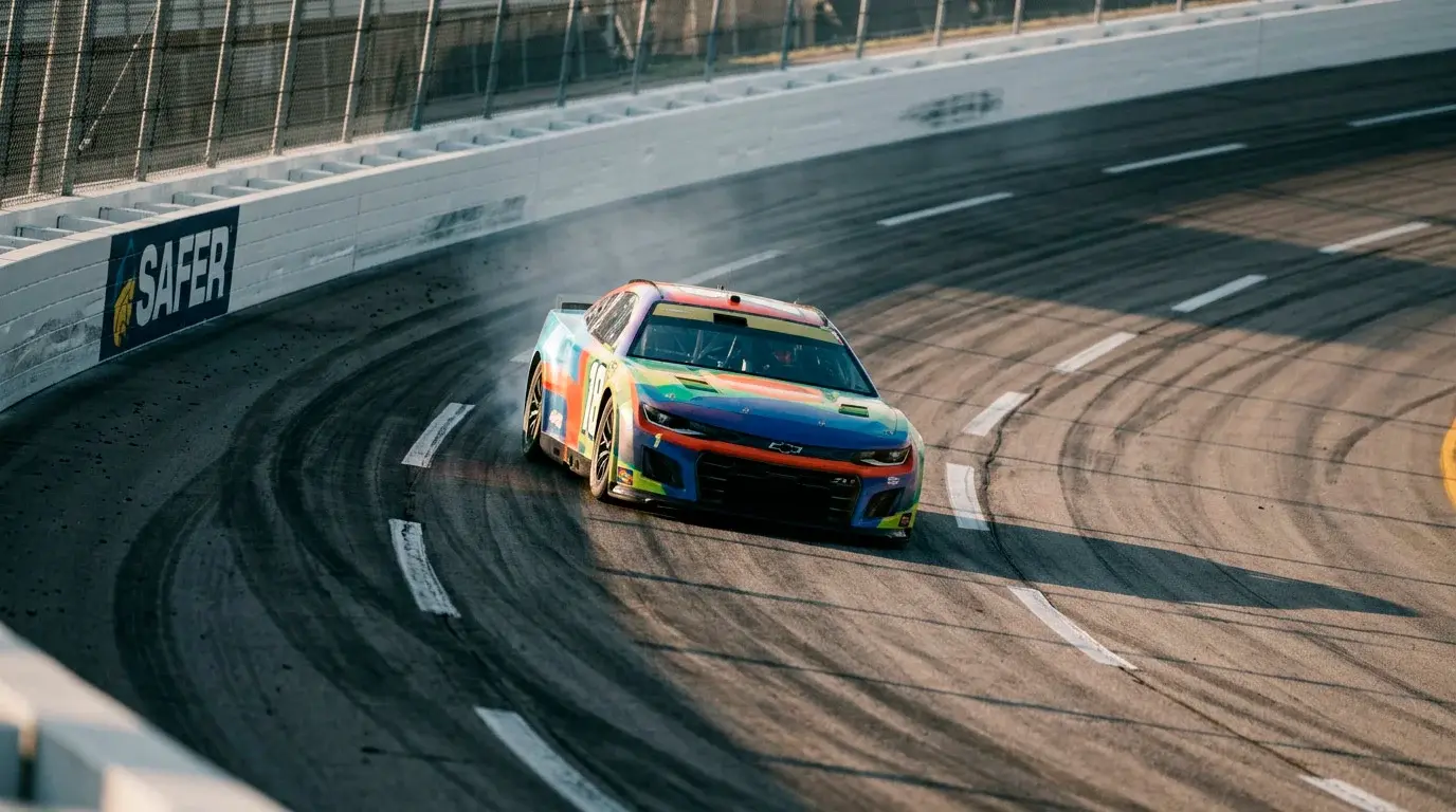 Coche NASCAR Cup Series tomando una curva peraltada de un óvalo de asfalto con marcas de neumáticos y muro SAFER al fondo